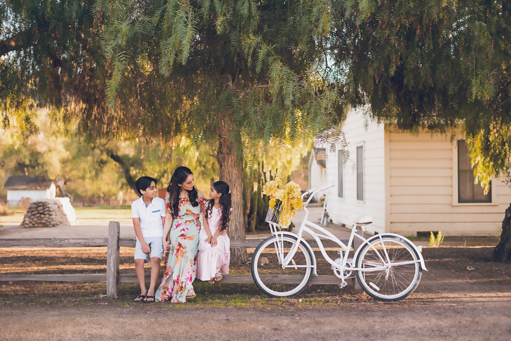 Mother, son and daughter sit on a wooden fence by a white bike with yellow flowers in a basket.