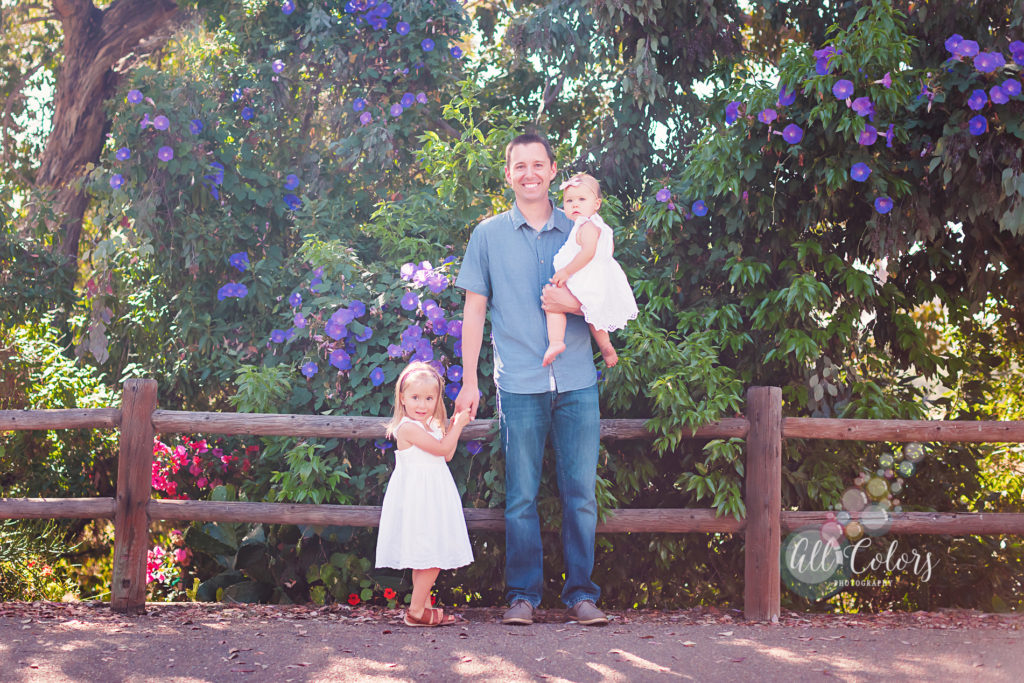 Father and two little girls in front of purple flower and a wooden fence.
