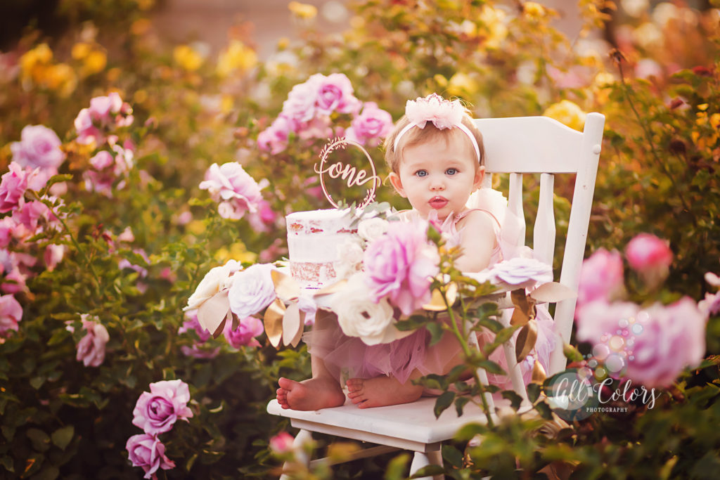 1 year old toddler sitting on a high chair eating cake on a rose garden.
