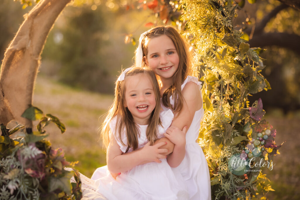 two sisters smiling and looking at the camera while sitting on a flower swing
